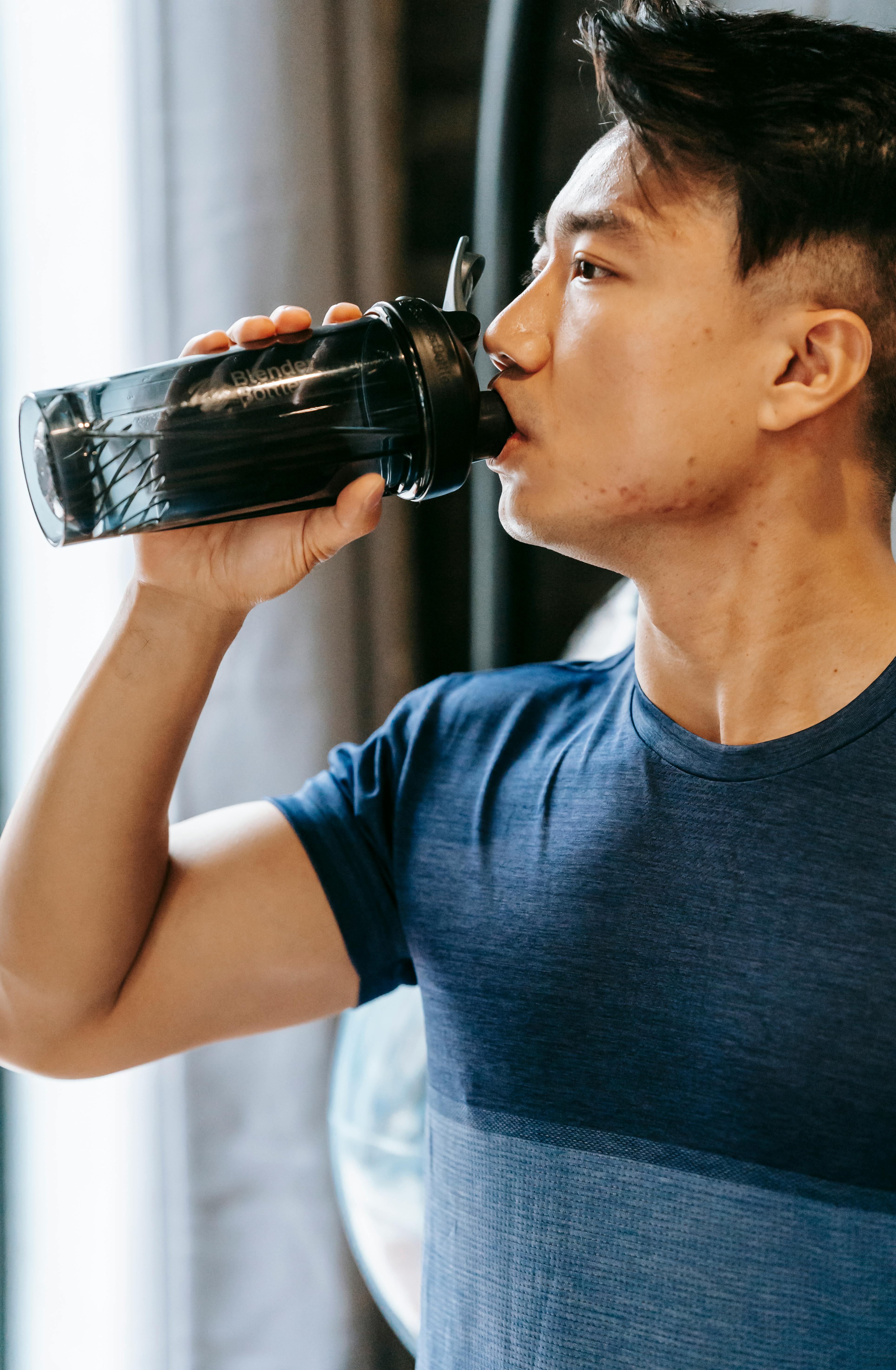 Serious ethnic man in sportswear drinking water from plastic bottle during break in fitness workout in gym